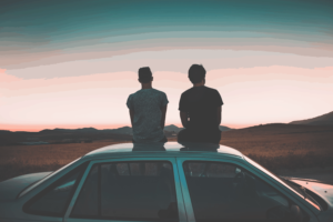 Two men sit on top of a car, facing away from the camera. They are looking towards a sunset.